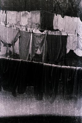 Vintage black-and-white photo of laundry hung on a communal line, featuring sheets, towels, and a large sack. The scene sugge...
