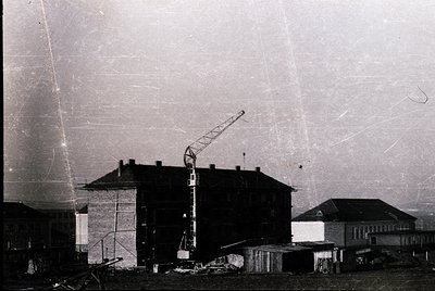 Mid-20th century construction site featuring a multi-story brick building under development. Tower crane positioned centrally...