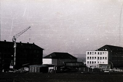 Black-and-white industrial site featuring mid-rise concrete buildings with dated "1971" marking on one structure. Crane and s...