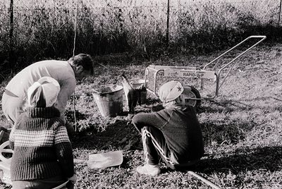 Two individuals in mid-20th-century outdoor attire (plaid shirt, sweater, headscarf) inspecting a buried metal container labe...