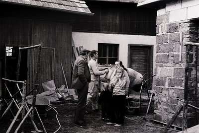 Three individuals in 1970s-era clothing gather in a rustic courtyard surrounded by wooden and stone structures. One man in a ...