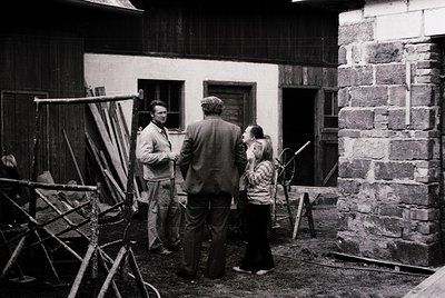 Three adults converse near a rustic stone-and-wood farmhouse, mid-20th century. Ladders, tools, and a wooden structure sugges...