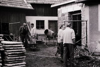 Four men in 1960s-70s workwear—hard hats, aprons, and rolled sleeves—engage in manual construction in a rural courtyard. Stac...