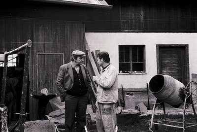 Two men inspecting a wooden beam in a construction site, likely mid-20th century. One wears a suit and cap, the other overall...