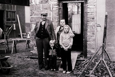 Family portrait outside a rustic brick home, likely mid-20th century. Four individuals pose in front of a weathered doorway w...
