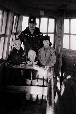 Family portrait in a rustic wooden cabin, likely mid-20th century. Adult male in uniform cap (likely military or scout) stand...