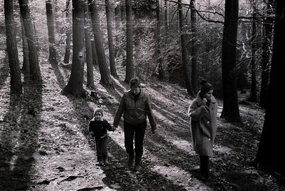Three figures in winter forest, mid-20th century attire. Adults in long coats, child in a knit hat and coat, walking on a sno...