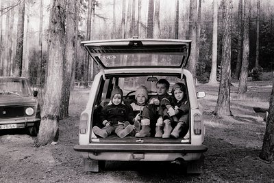 Four children sit in an open rear hatch of a vintage station wagon, surrounded by a forest of tall, leafless trees. Mid-20th ...