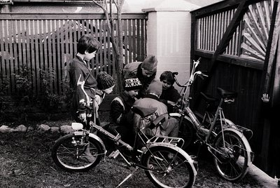 Four children in 1970s-era streetwear gather around vintage BMX-style bikes in a residential courtyard. Wooden fence and bric...