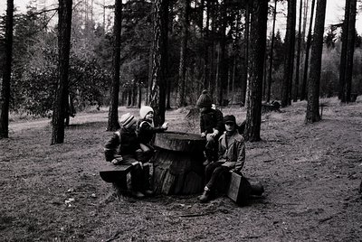 Three individuals seated on a tree stump in a dense forest, mid-20th century. Traditional rural attire suggests agricultural ...