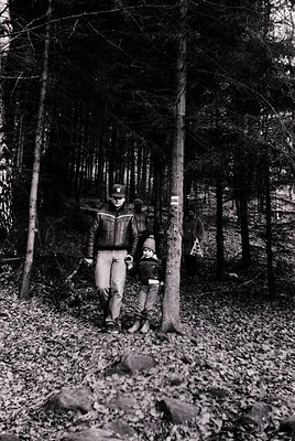 Black-and-white forest scene featuring a man and child hiking on a dirt trail. The man wears a striped sweater, cap, and trou...