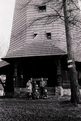Family portrait in front of a rustic wooden tower house, likely mid-20th century. Five individuals pose on stone steps, dress...