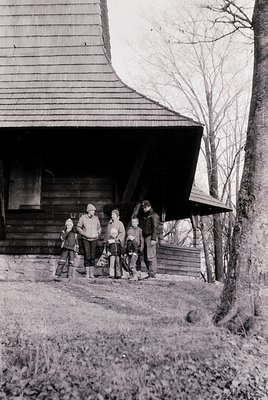 Family group poses outside rustic wooden cabin with sloped roof, likely mid-20th century. Adults in heavy coats, children in ...