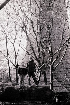 Two children pose on a stone ledge in winter attire—knit hats, jackets, and boots—against a bare tree and brick wall. Likely ...