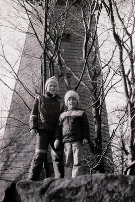 Two individuals pose outdoors in winter attire against a tall, cylindrical brick tower with arched windows. The adult wears a...