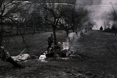 Two men in mid-20th-century workwear tend a smoldering fire in a barren field, surrounded by leafless trees. One kneels, shov...