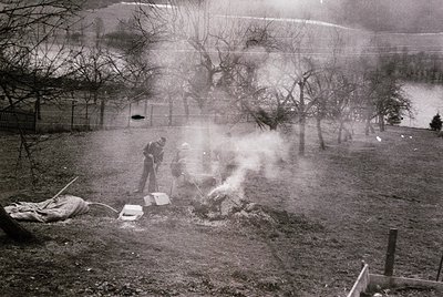 Mid-20th century black-and-white photo of two men tending a bonfire in a rural backyard. One man stokes flames with a shovel;...