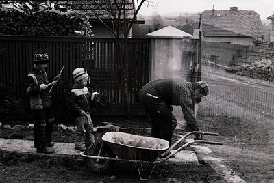 Three construction workers in mid-20th-century attire—hard hats, striped shirts, and aprons—operate near a residential fence....