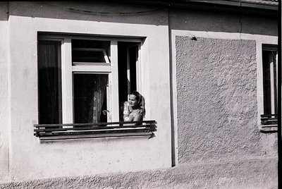 A woman leans from a window with a wrought-iron balcony, dressed in a sleeveless top and skirt, set in a weathered plaster bu...