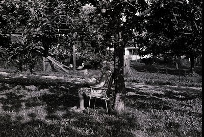 Mid-century black-and-white photo of a woman reading in a garden chair under leafy trees, with a vintage hammock and wooden f...