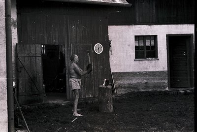 Black-and-white rural scene: elderly man in minimalist loincloth wielding a hammer near weathered wooden barn door with targe...