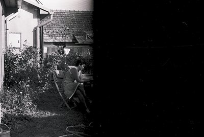 Vintage black-and-white photo of a woman seated on a wicker chair in a sunlit courtyard, reading or writing. Wooden house wit...