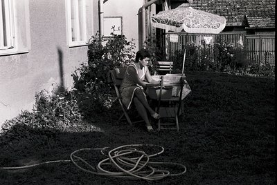 A woman in a floral dress sits on a wicker patio chair, reading outdoors in a mid-20th-century residential courtyard. Decorat...