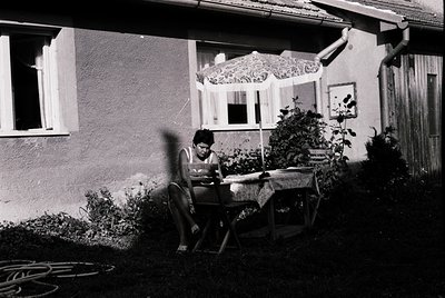 A woman sits at a small wooden table outdoors, reading a book under a decorative lace curtain. The setting appears to be a re...