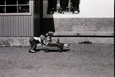 Two children in mid-20th-century clothing push a toy wagon on a gravel driveway beside a brick house with wooden shutters. Cl...