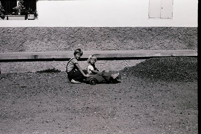 Vintage black-and-white photo of two children playing on a dirt surface beside a concrete wall. Older child pushes younger ch...