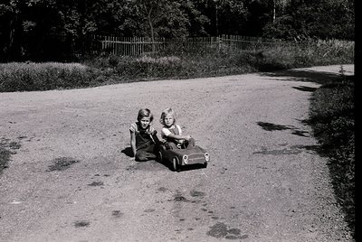 Two young girls in vintage-style dresses play with a toy car on a gravel path, framed by overgrown greenery and a wooden fenc...