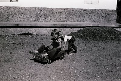 Mid-20th century black-and-white photo of a child riding a homemade cart on a rough concrete surface. Child wears a striped s...