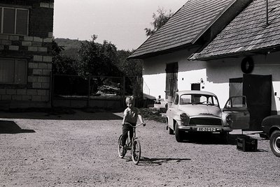 A young boy rides a simple bicycle in a gravel courtyard beside a vintage car ( ).