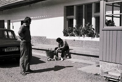 Black-and-white snapshot of two men in 1970s Alpine village. One kneels tending to a dog in a basket beside a vintage car (li...