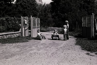 Vintage black-and-white photo of three children playing in a park playground, mid-20th century. Two boys sit on a small bench...