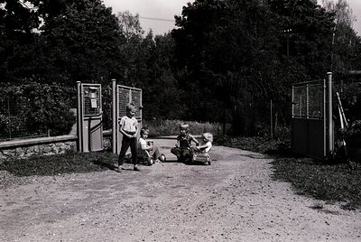 Vintage black-and-white photo of three children playing with a scooter on a gravel path beside public phone booths, likely mi...