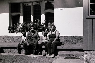 Mid-20th century family portrait on a sunlit porch with potted plants. Three adults and two children pose on stone steps, dre...
