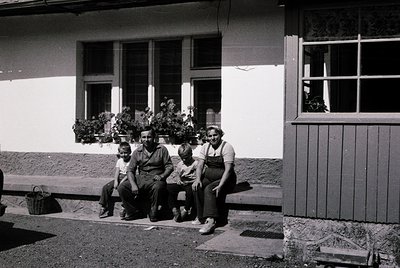 Family portrait in front of a modest, single-story house with floral window boxes, mid-20th century. Three adults and two chi...