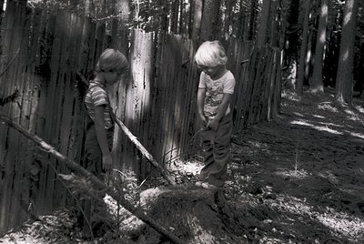 Two children in 1970s-style clothing play with wooden planks in a forested area. One holds a shovel, the other a stick, near ...