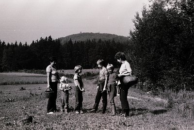 Five adults and children in rural field, mid-20th century. Group poses with woven baskets, likely foraging or picnicking. Lus...