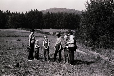 A mid-20th century rural gathering in a lush, forested landscape. Six children and two adults pose on a grassy hillside, fram...
