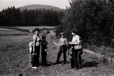 Four adults and children pose outdoors in a rural setting, likely mid-20th century. Two women stand on right holding baskets;...