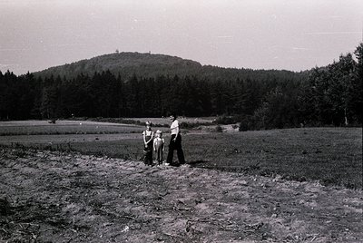 Mid-20th century rural family portrait in a plowed field with forested hillside backdrop. Three adults and two children pose ...