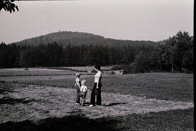 Three figures stand on a grassy hillside overlooking a forested ridge, mid-20th century. Adult in long-sleeve shirt and trous...
