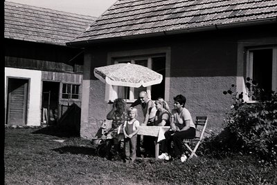 Mid-century family portrait in a residential courtyard, featuring a light blue stucco house with wooden accents. Four adults ...