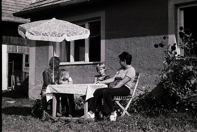 Family gathers under a floral-patterned patio umbrella at a mid-century residential table, set with a white cloth. Two adults...