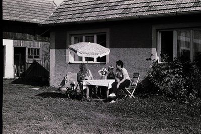 Mid-century outdoor scene featuring three adults seated at a small wooden table under a striped patio umbrella. The house dis...