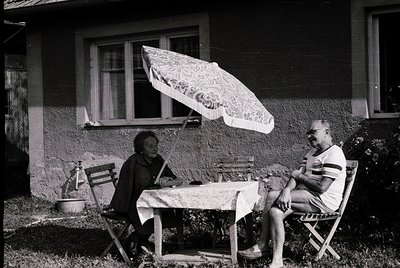 Two individuals seated outdoors under a floral-patterned umbrella, 1960s-1970s. Wooden chairs and table with a white tableclo...