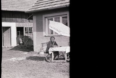 Mid-20th century rural home exterior with a woman seated on a folding chair, ironing near a wooden door. House features simpl...