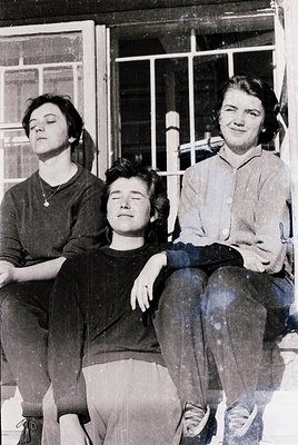 Three women pose casually in a mid-20th-century setting, likely 1950s–1960s. Left wears a fitted sweater with a pendant neckl...